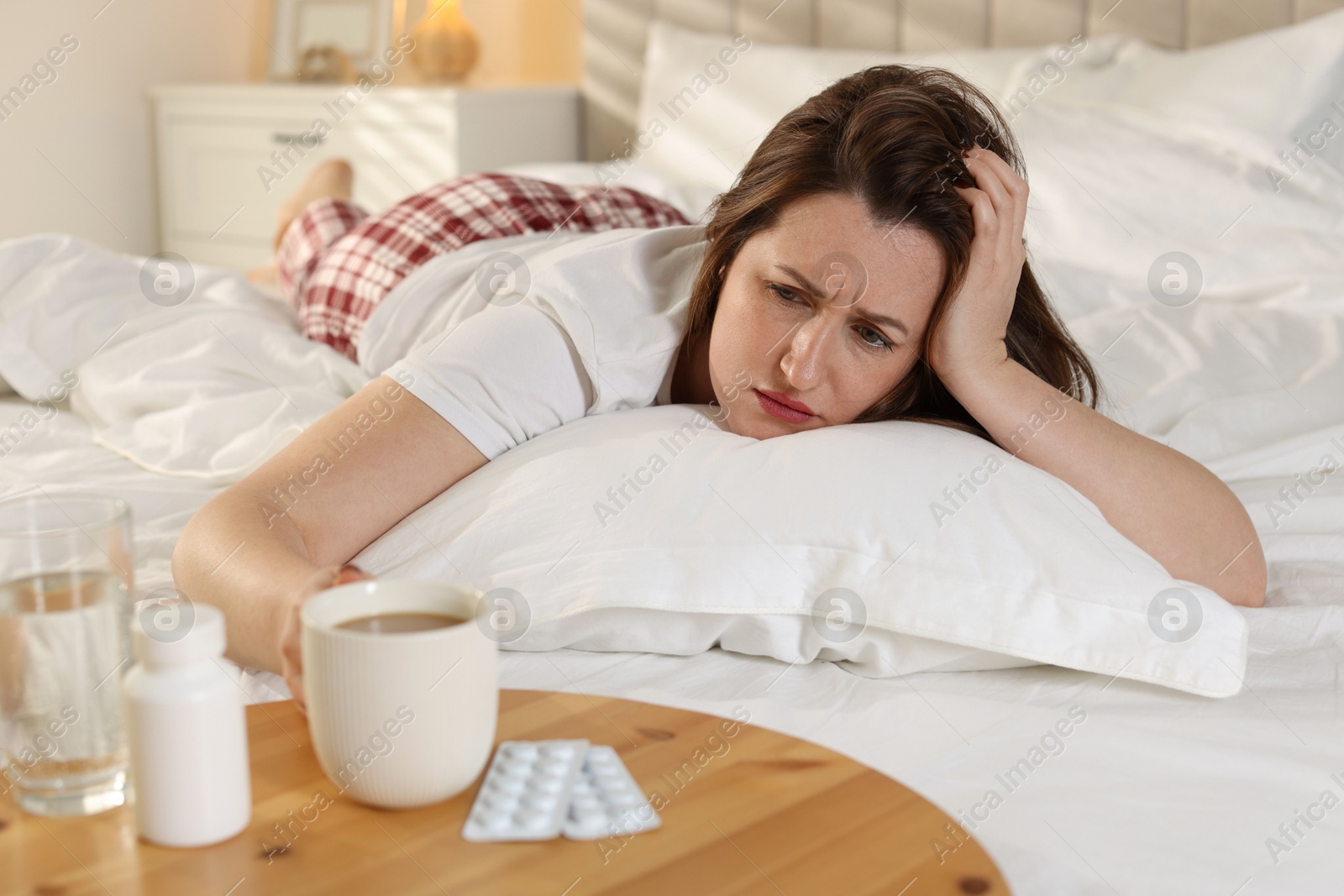 Photo of Unhappy woman with cup of coffee, water and pills suffering from hangover on bed at home