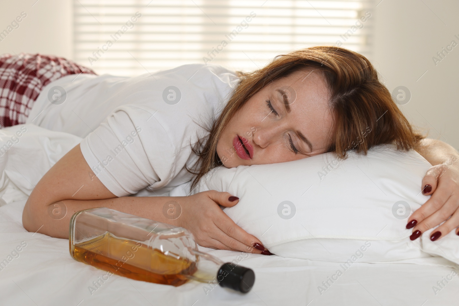 Photo of Suffering from hangover. Woman with bottle of whiskey sleeping on bed at home