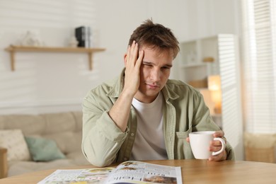 Photo of Suffering from hangover. Unhappy young man with cup of coffee sitting at wooden table indoors