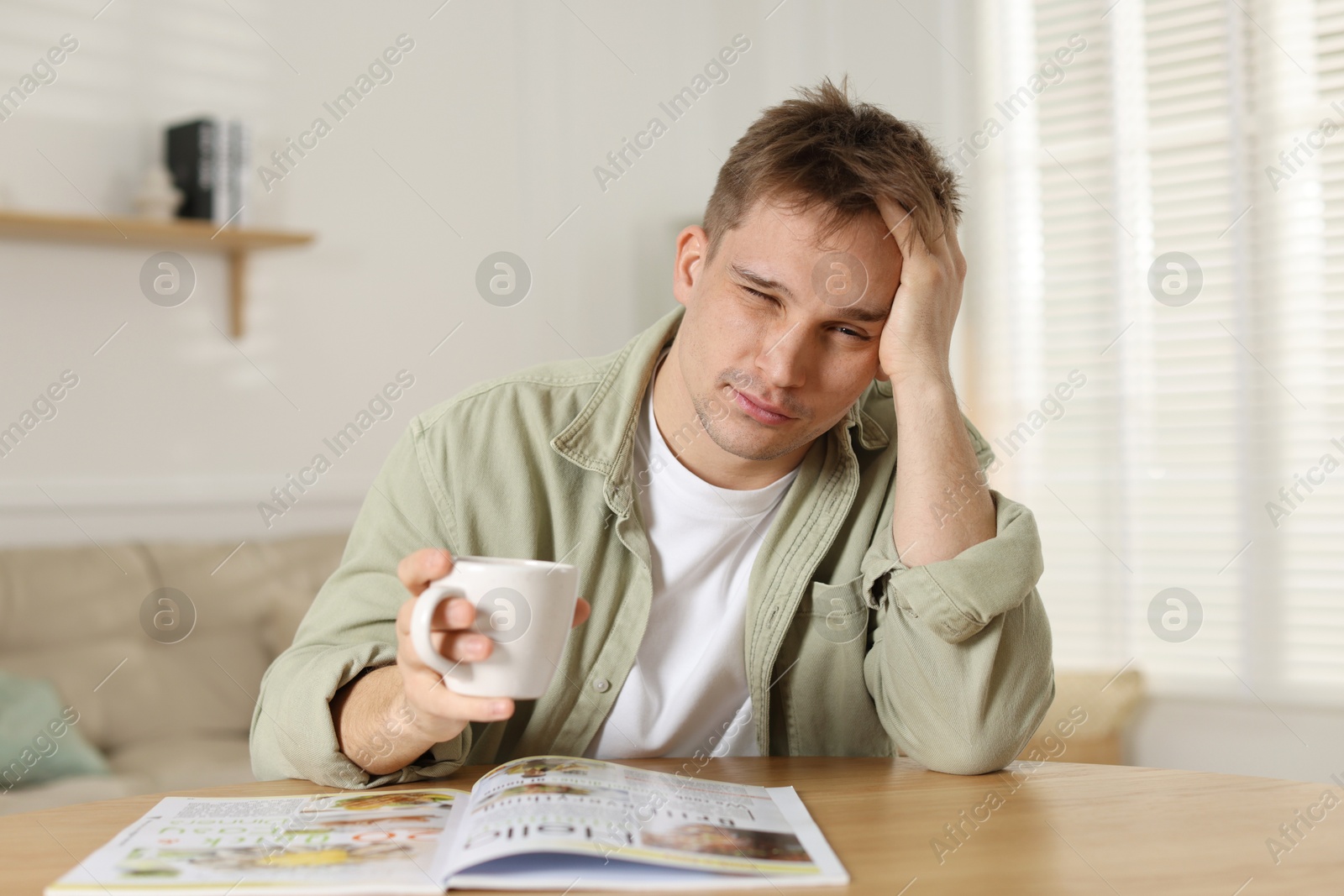 Photo of Suffering from hangover. Unhappy young man with cup of coffee sitting at wooden table indoors