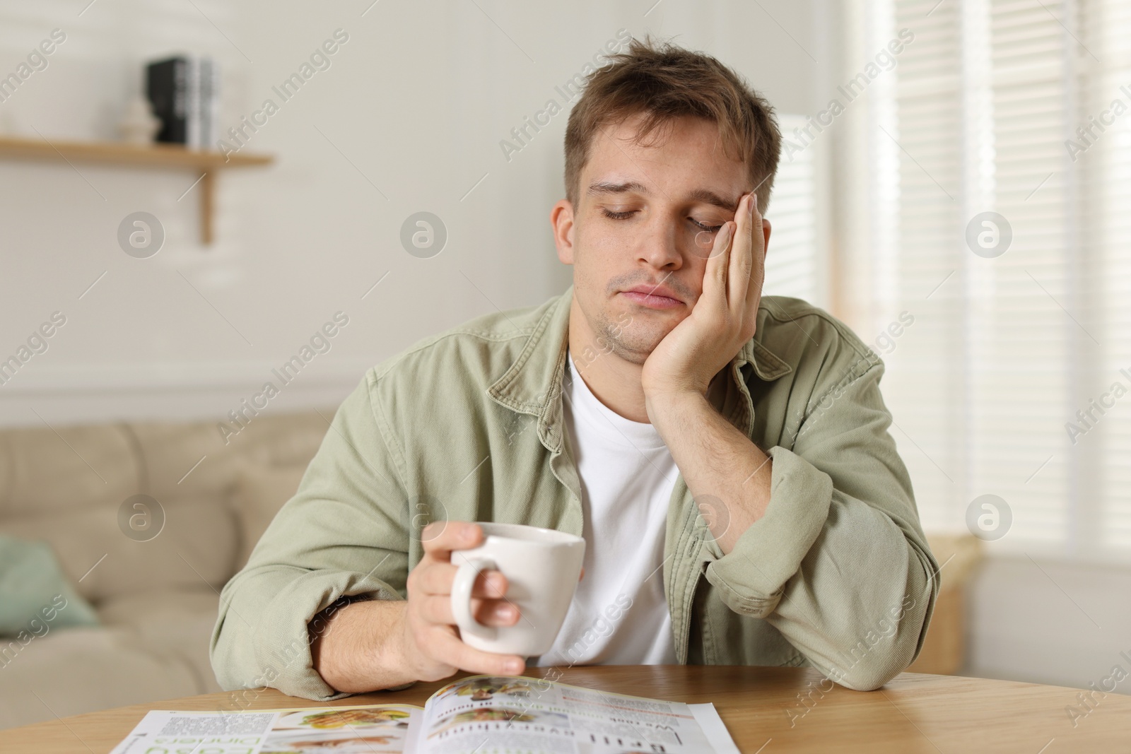 Photo of Suffering from hangover. Unhappy young man with cup of coffee sitting at wooden table indoors