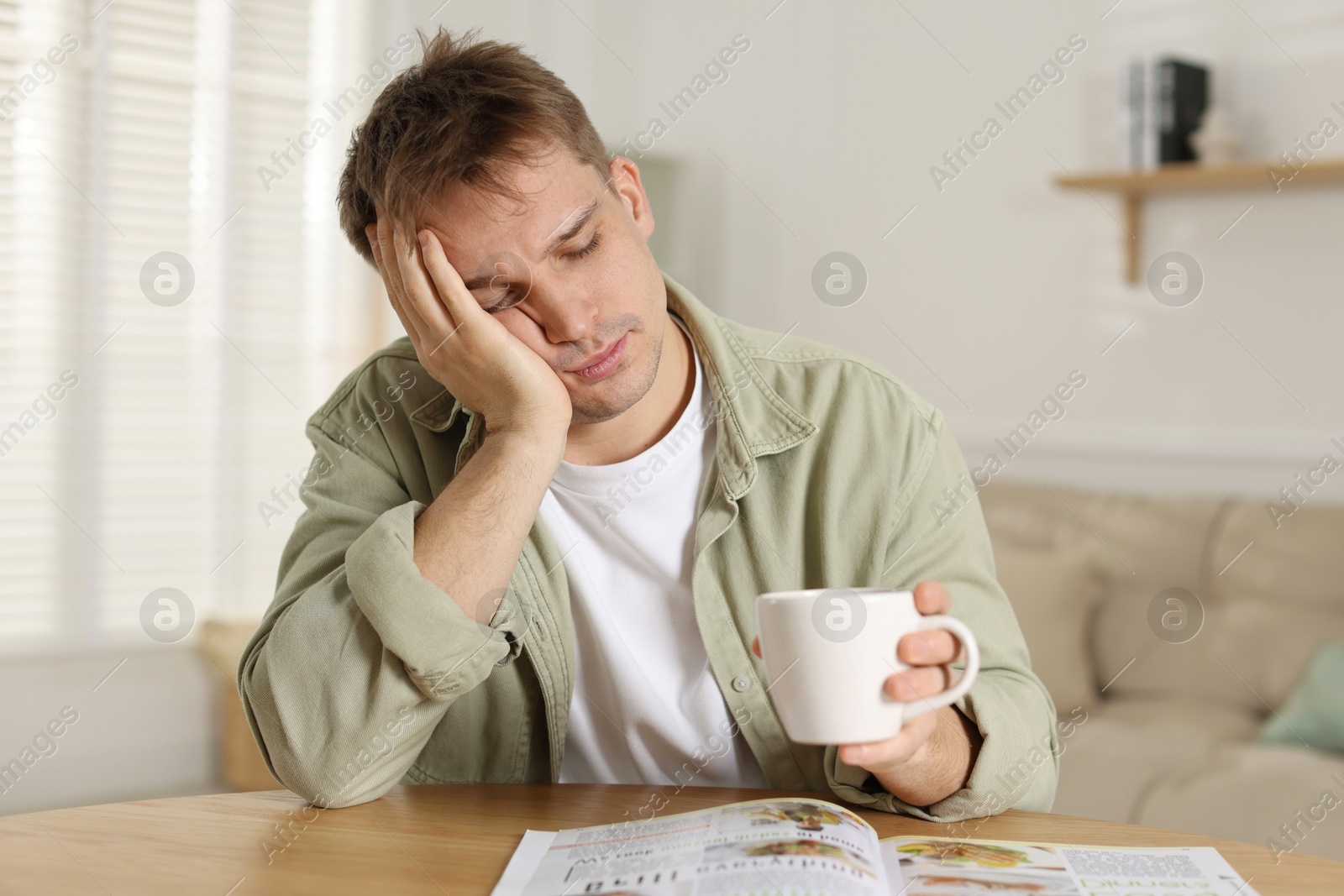 Photo of Suffering from hangover. Unhappy young man with cup of coffee sitting at wooden table indoors