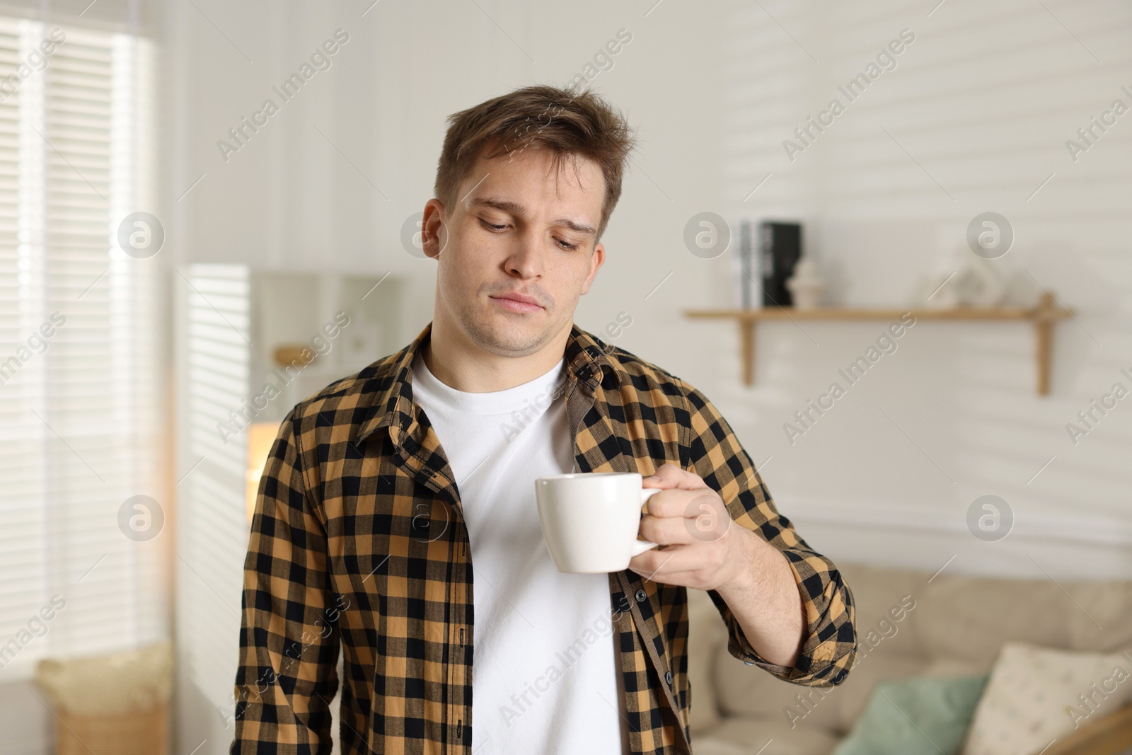 Photo of Unhappy young man with cup of coffee suffering from hangover at home