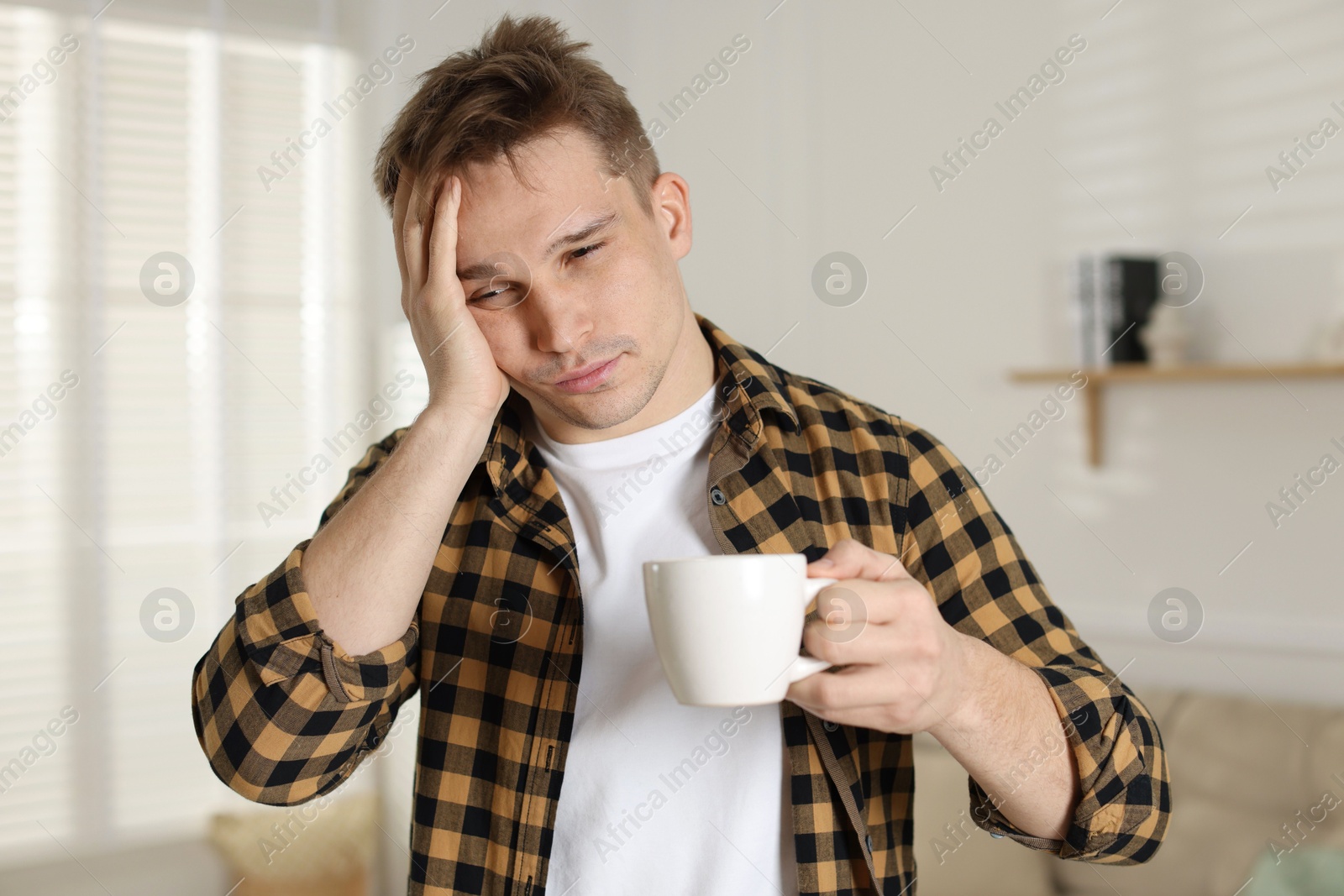 Photo of Unhappy young man with cup of coffee suffering from hangover at home