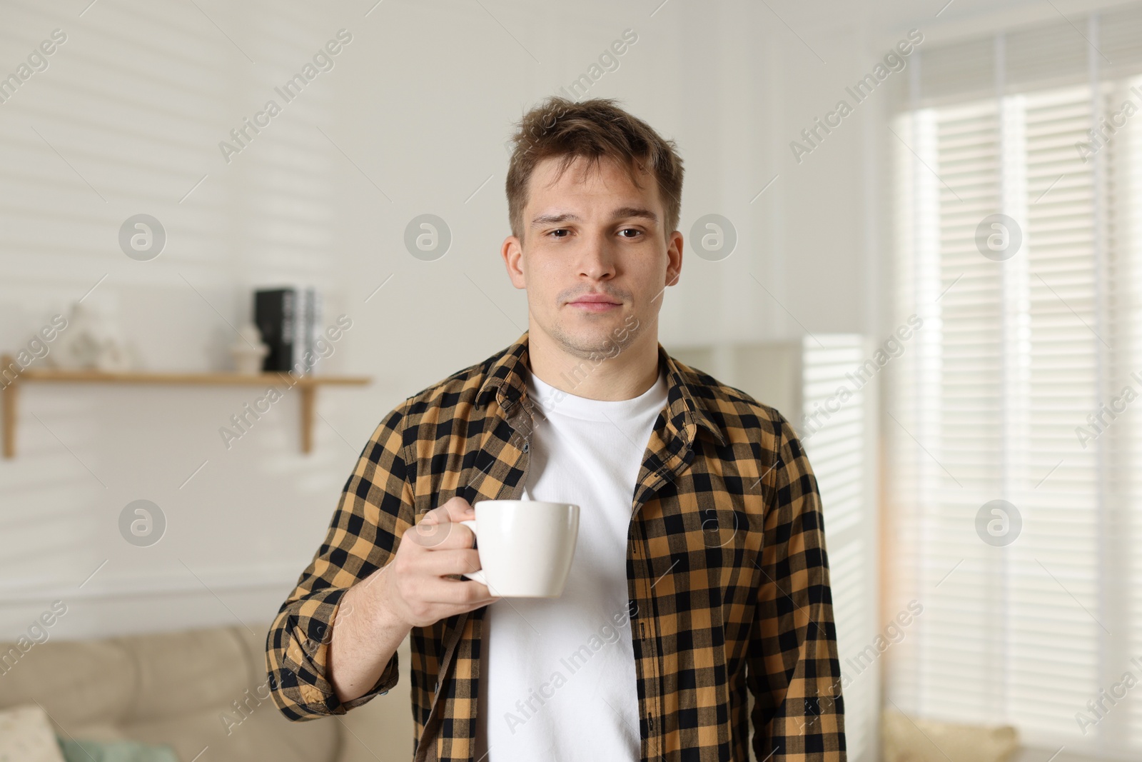 Photo of Unhappy young man with cup of coffee suffering from hangover at home