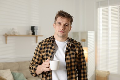 Unhappy young man with cup of coffee suffering from hangover at home