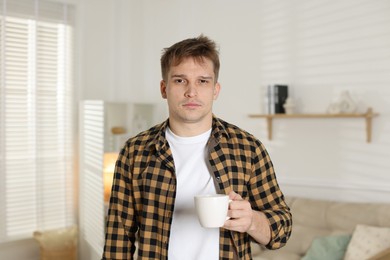 Unhappy young man with cup of coffee suffering from hangover at home