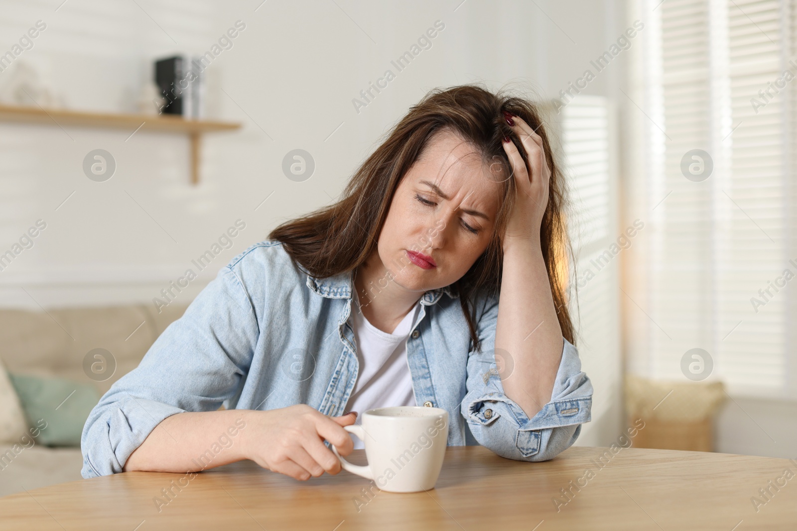 Photo of Suffering from hangover. Unhappy woman with cup of coffee sitting at wooden table indoors