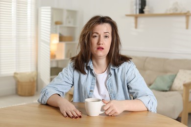 Suffering from hangover. Unhappy woman with cup of coffee sitting at wooden table indoors