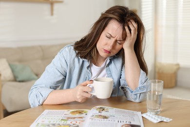 Suffering from hangover. Unhappy woman with cup of coffee sitting at wooden table indoors