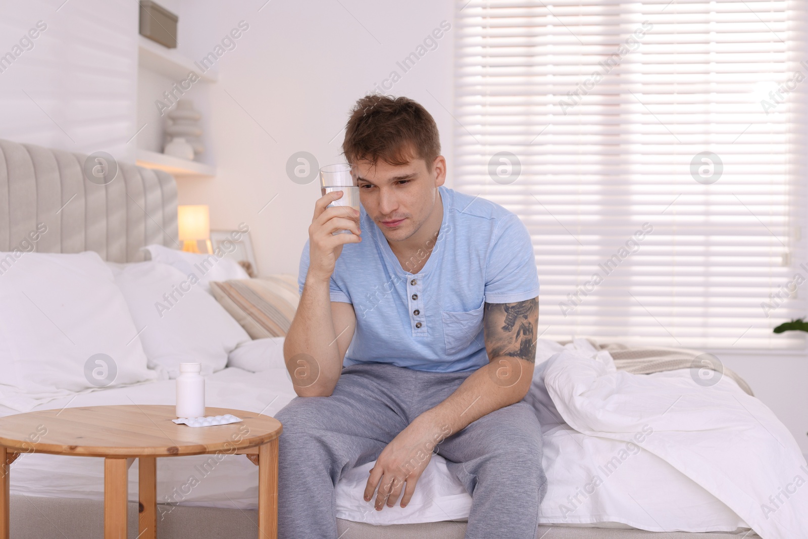 Photo of Unhappy young man with glass of water suffering from hangover on bed at home