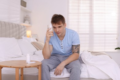 Unhappy young man with glass of water suffering from hangover on bed at home