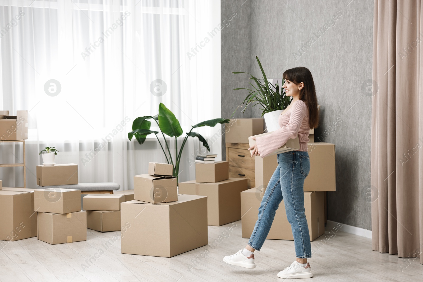 Photo of Moving day. Happy woman with cardboard box and houseplant in her new home