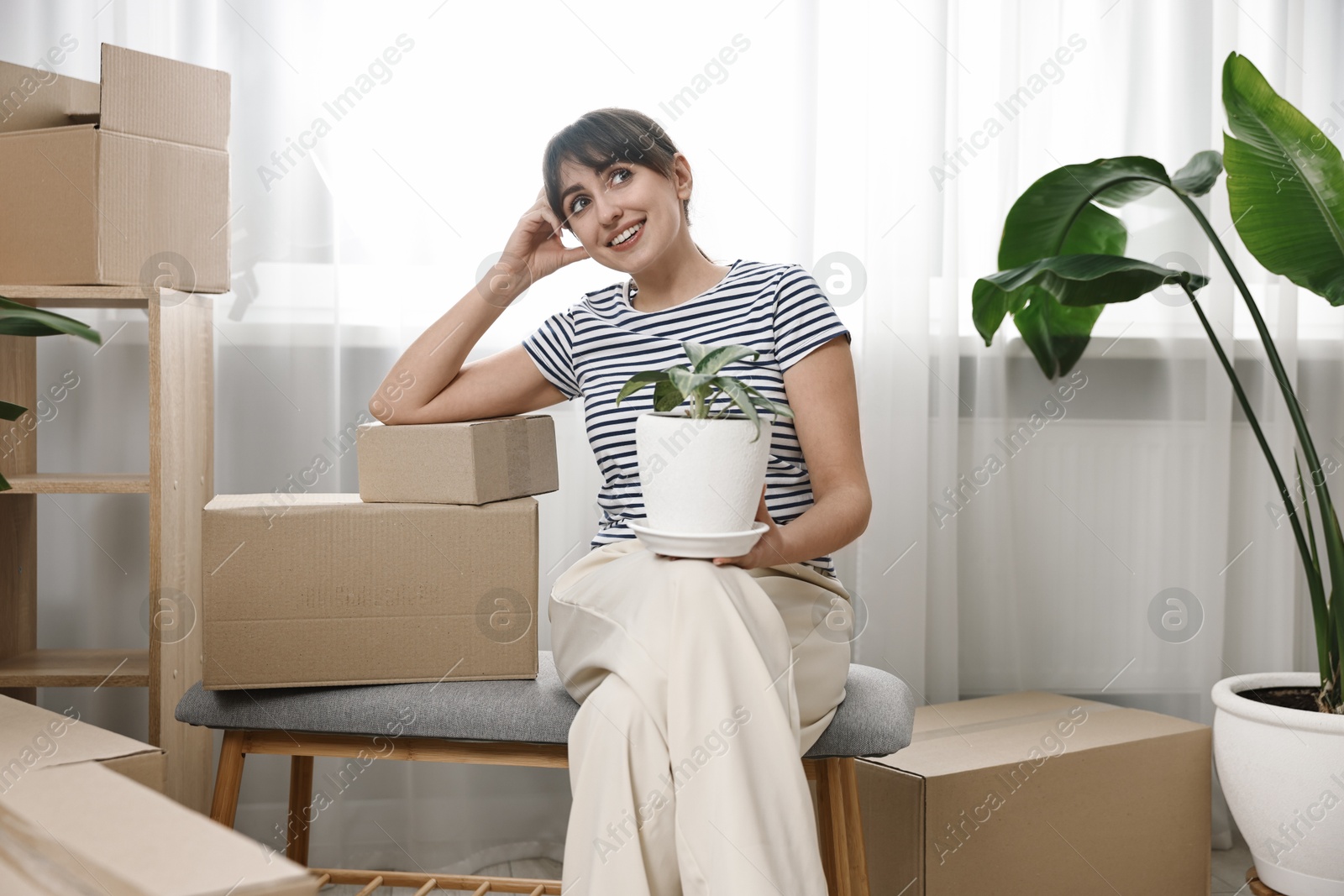 Photo of Moving day. Happy woman with houseplant and cardboard boxes in her new home