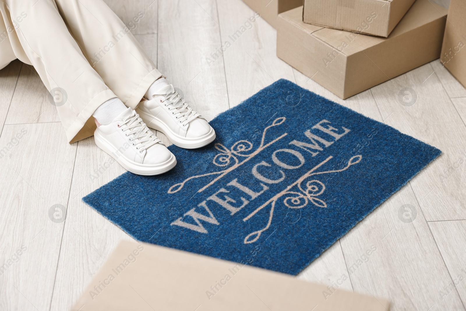 Photo of Moving day. Woman sitting near cardboard boxes and doormat with word Welcome on floor in her new home, closeup