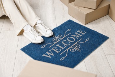 Photo of Moving day. Woman sitting near cardboard boxes and doormat with word Welcome on floor in her new home, closeup