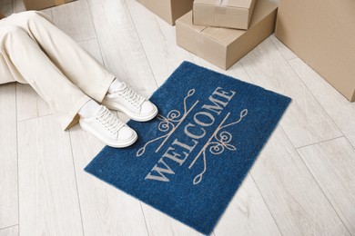 Photo of Moving day. Woman sitting near cardboard boxes and doormat with word Welcome on floor in her new home, closeup