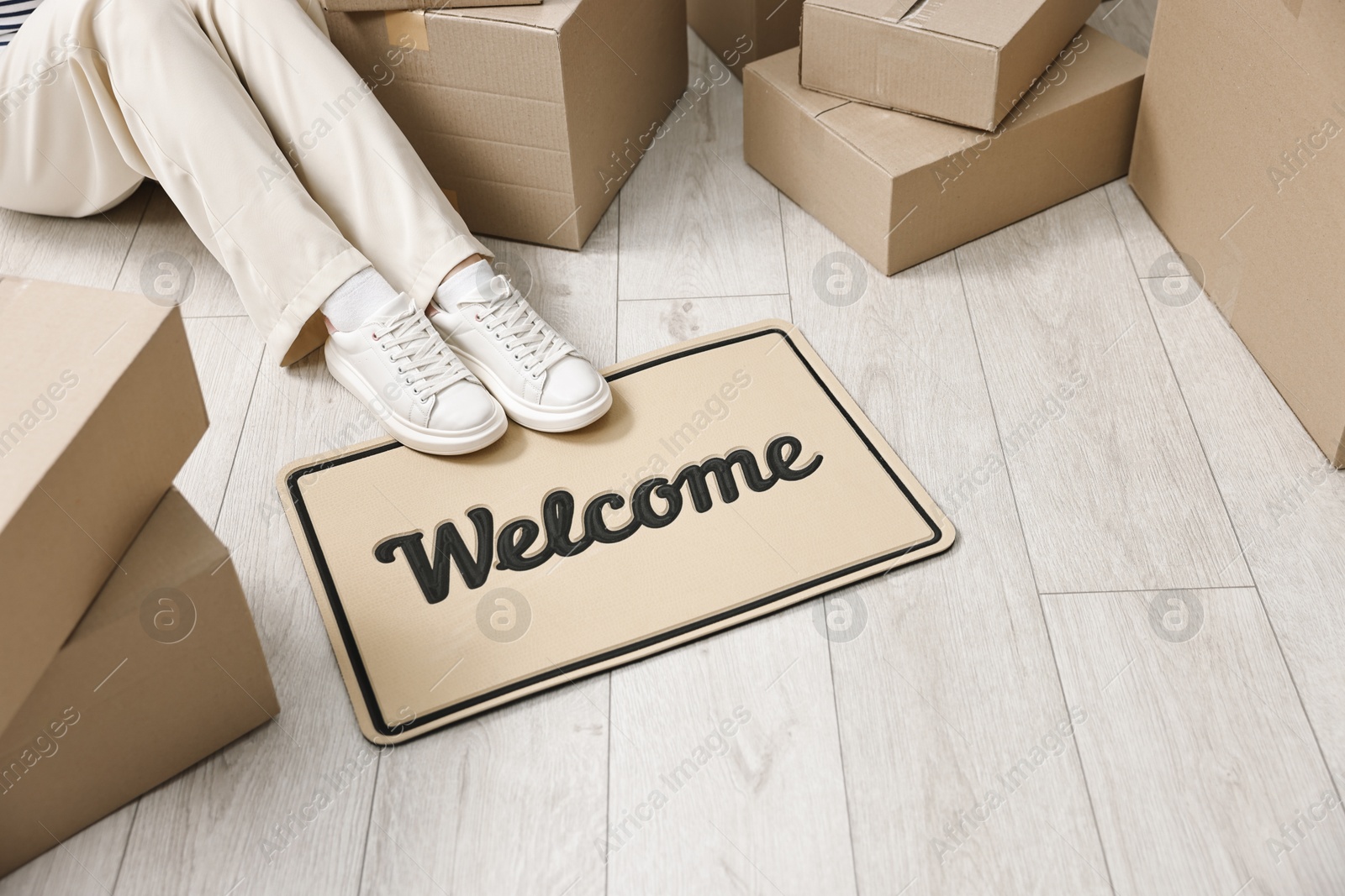 Photo of Moving day. Woman sitting near cardboard boxes and doormat with word Welcome on floor in her new home, closeup