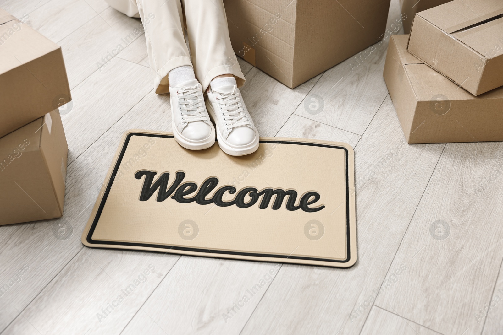 Photo of Moving day. Woman sitting near cardboard boxes and doormat with word Welcome on floor in her new home, closeup