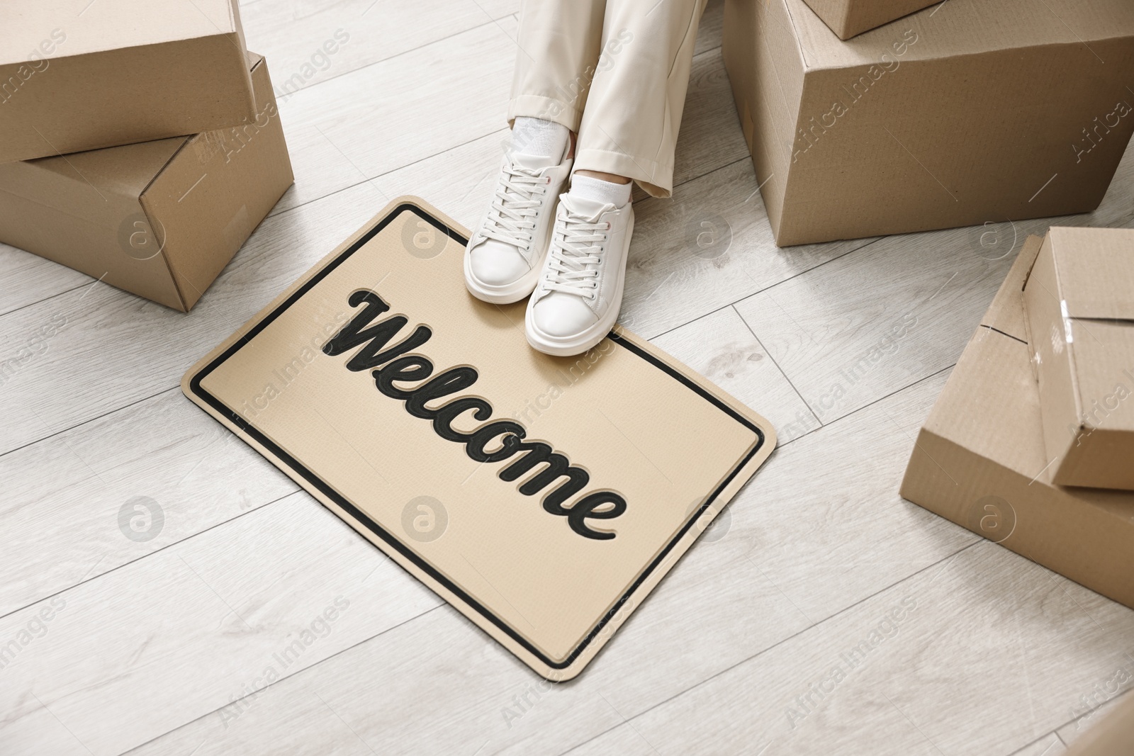 Photo of Moving day. Woman sitting near cardboard boxes and doormat with word Welcome on floor in her new home, closeup