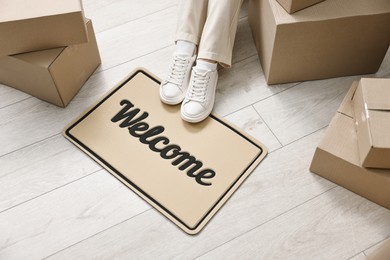 Photo of Moving day. Woman sitting near cardboard boxes and doormat with word Welcome on floor in her new home, closeup