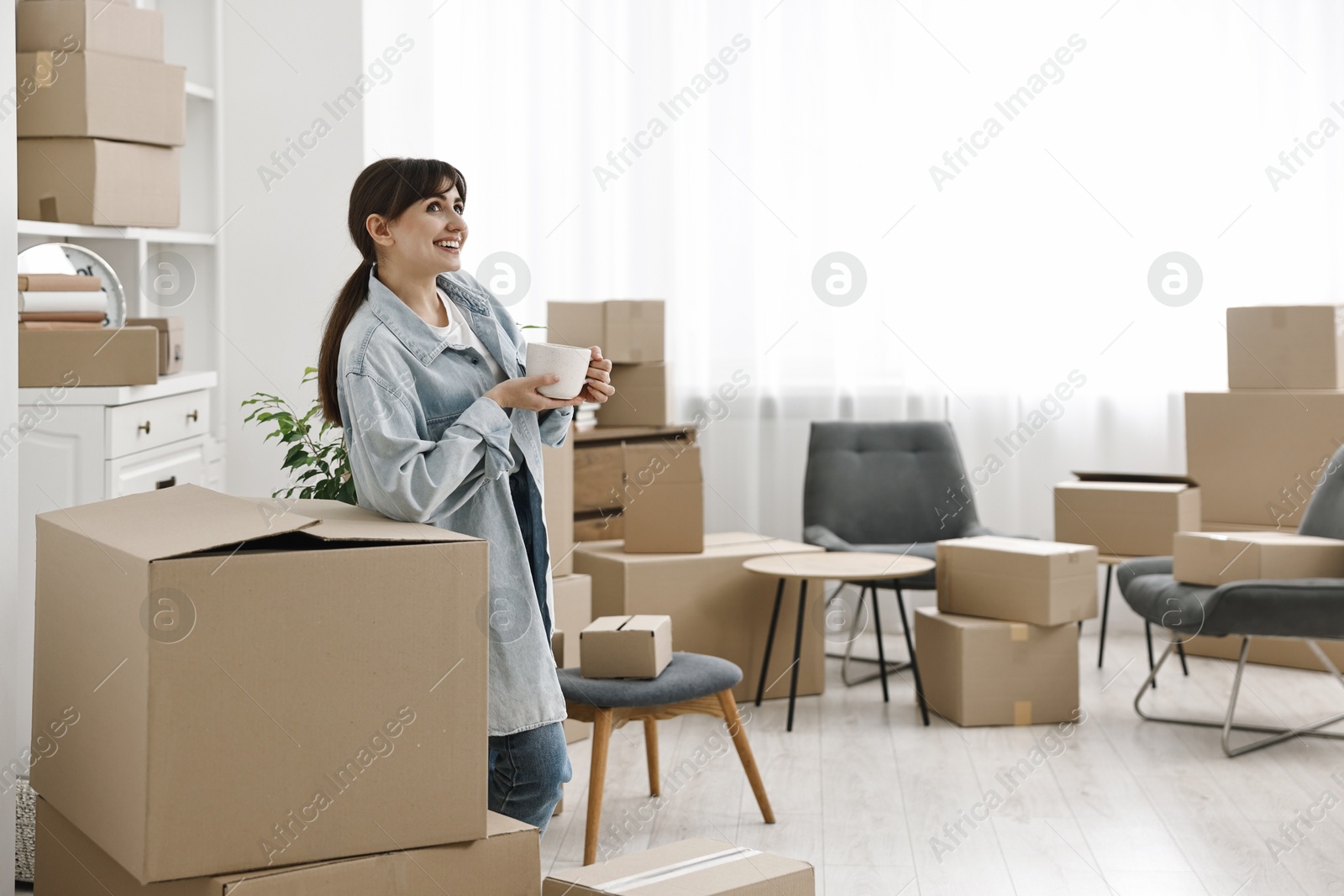 Photo of Moving day. Happy woman with cup of drink and cardboard boxes in her new home