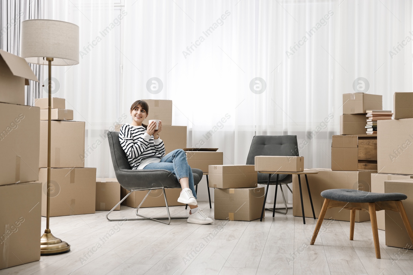 Photo of Moving day. Happy woman with cup of drink and cardboard boxes in new home