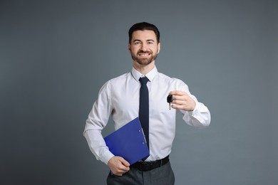 Cheerful salesman with car key and clipboard on grey background