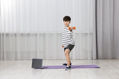 Photo of Little boy exercising with dumbbells near laptop at home. Morning routine