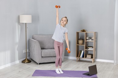 Photo of Little girl exercising with dumbbells near laptop at home. Morning routine