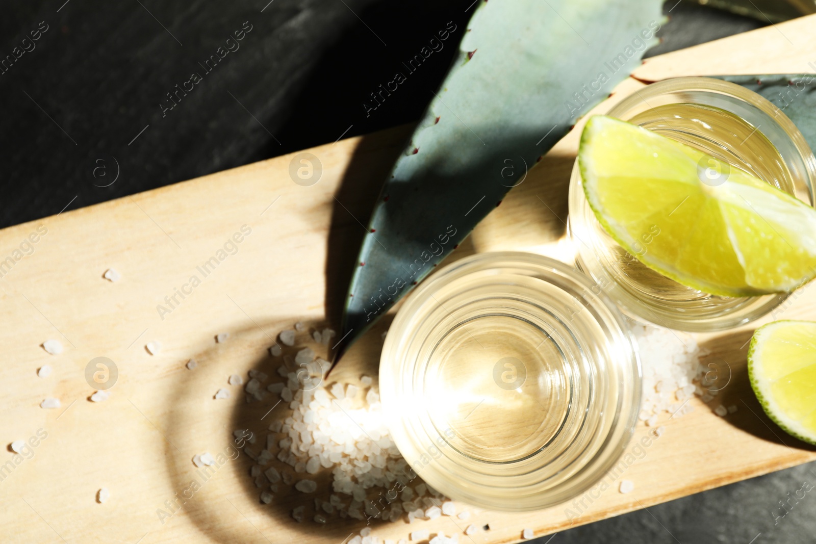 Photo of Tequila shots, slices of lime, salt and agave leaves on dark table, flat lay