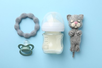 Feeding bottle with milk, needle felted cat, teether and pacifier on light blue background, flat lay