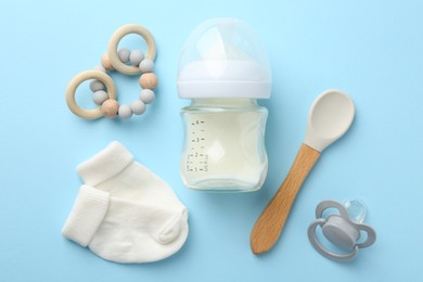 Feeding bottle with milk, baby socks, spoon, teether and pacifier on light blue background, flat lay