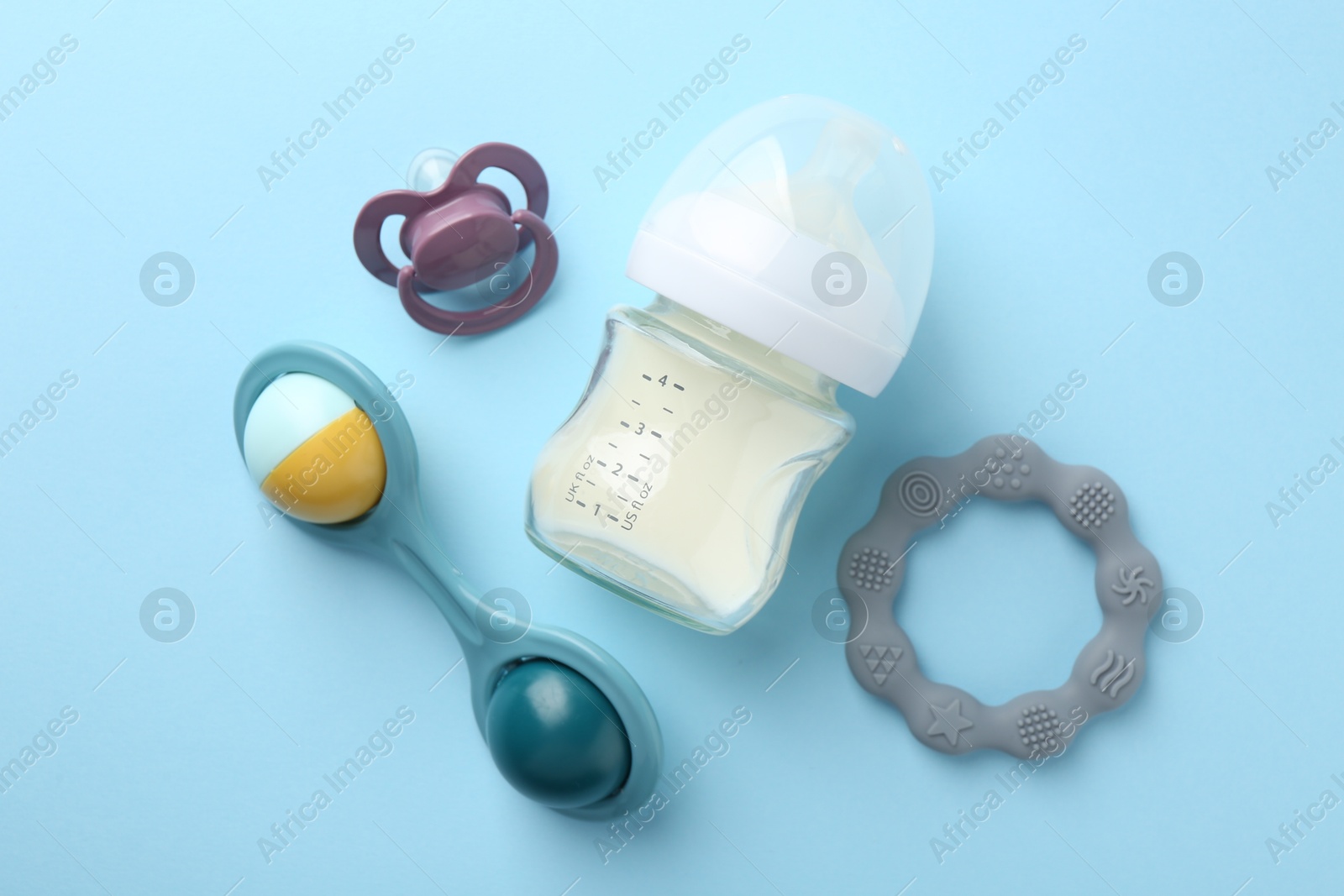 Photo of Feeding bottle with milk, baby rattle, teether and pacifier on light blue background, flat lay