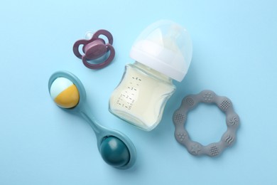 Feeding bottle with milk, baby rattle, teether and pacifier on light blue background, flat lay