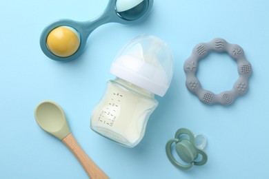 Photo of Feeding bottle with milk, baby rattle, teether, spoon and pacifier on light blue background, flat lay