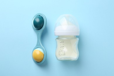 Feeding bottle with milk and baby rattle on light blue background, flat lay