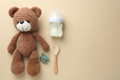 Feeding bottle with milk, toy bear, pacifier and spoon on beige background, flat lay. Space for text