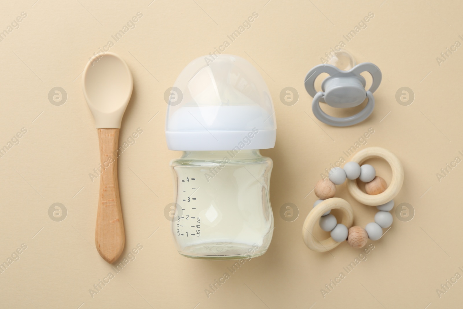 Photo of Feeding bottle with milk, pacifier, teether and spoon on beige background, flat lay