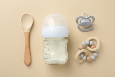 Feeding bottle with milk, pacifier, teether and spoon on beige background, flat lay