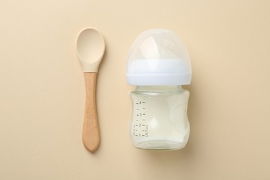 Feeding bottle with milk and spoon on beige background, flat lay