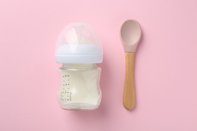 Feeding bottle with milk and spoon on pink background, flat lay