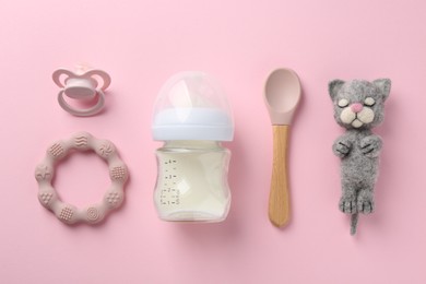 Feeding bottle with milk, needle felted cat, spoon, pacifier and teether on pink background, flat lay