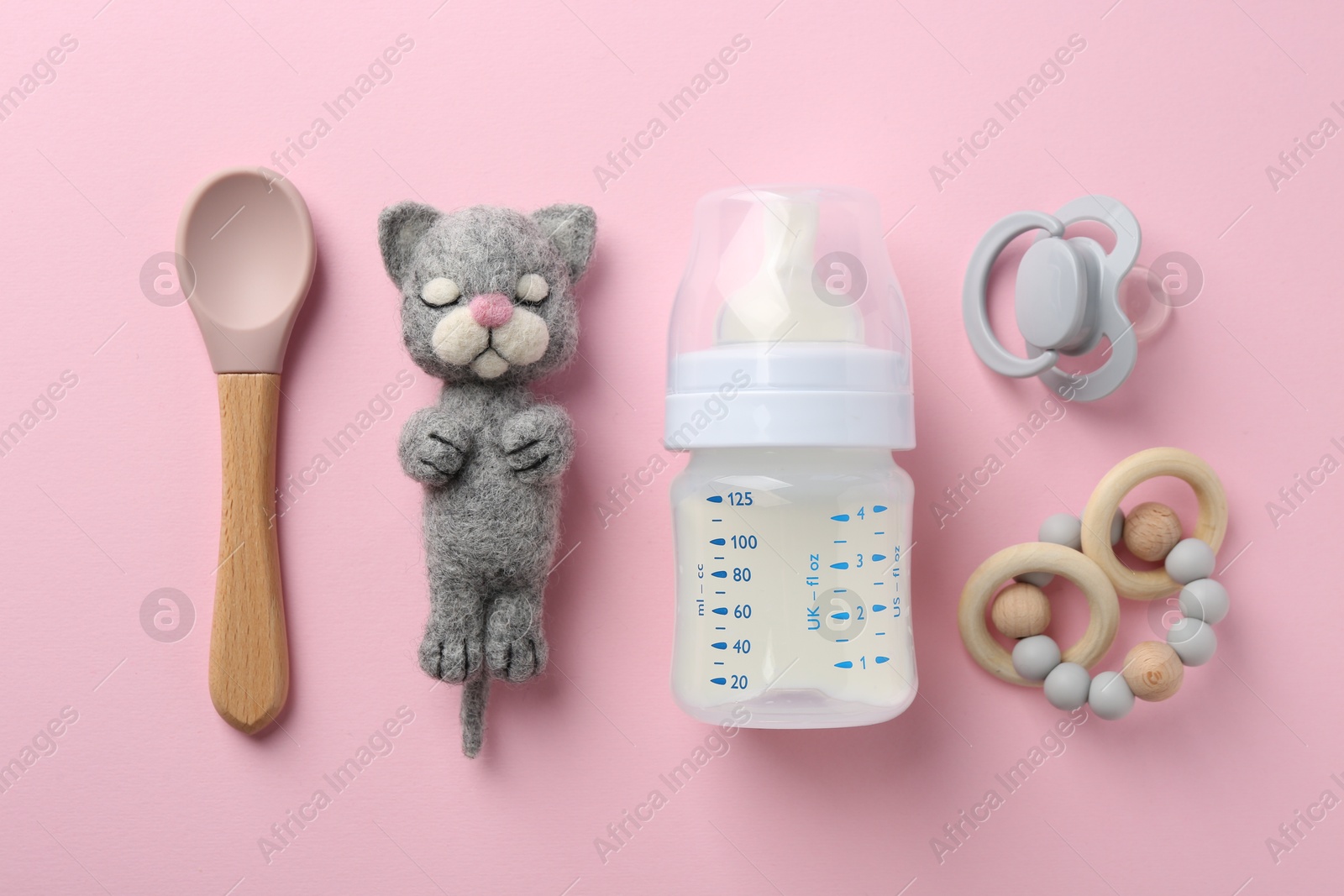 Photo of Feeding bottle with milk, needle felted cat, spoon, pacifier and teether on pink background, flat lay