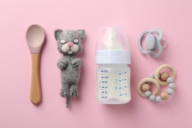 Feeding bottle with milk, needle felted cat, spoon, pacifier and teether on pink background, flat lay