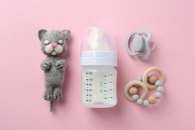 Feeding bottle with milk, needle felted cat, pacifier and teether on pink background, flat lay