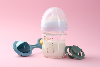 Feeding bottle with milk, pacifier and baby rattle on pink background