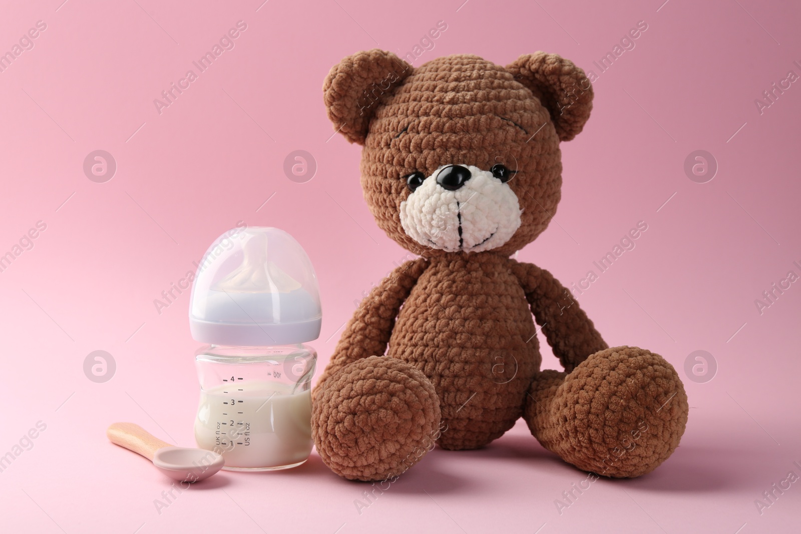 Photo of Feeding bottle with milk, spoon and toy bear on pink background