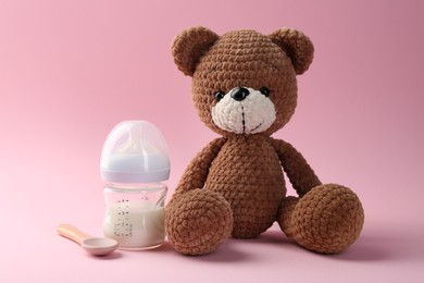 Photo of Feeding bottle with milk, spoon and toy bear on pink background