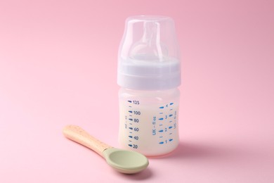 Feeding bottle with milk and spoon on pink background